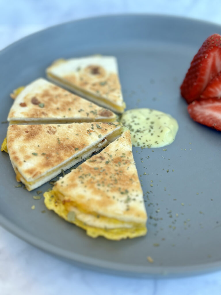 Wedges of an egg, cheese, and avocado quesadilla, sided with an avocado yogurt sauce and strawberries.