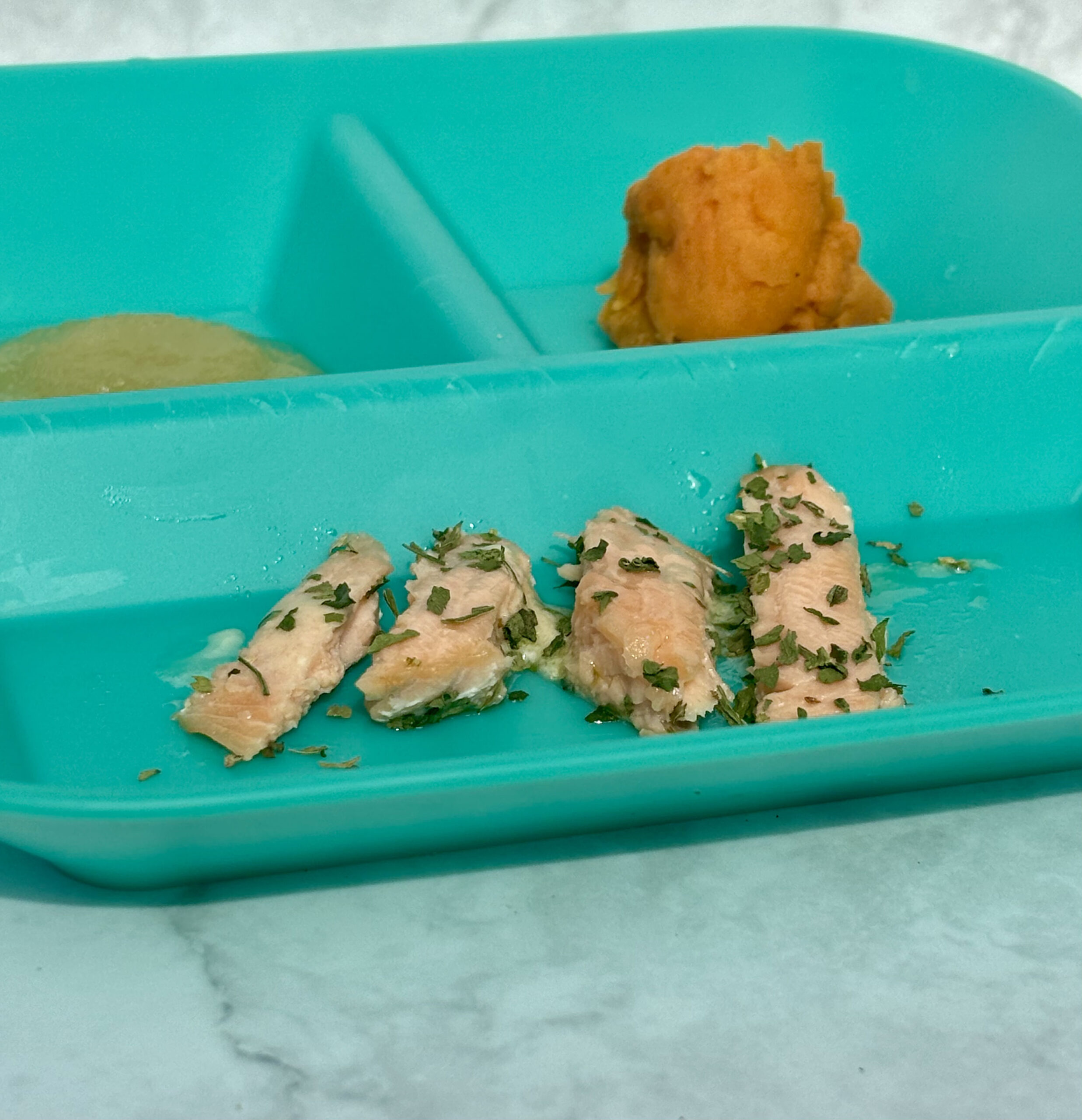 A plate with nuggets of salmon; applesauce and mashed sweet potatoes appear in the background.