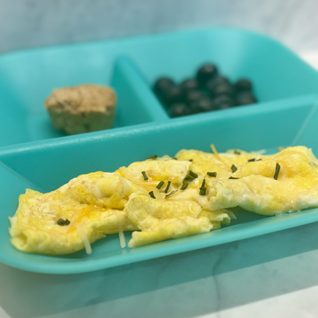 A crepe style omelette that's the right size and texture for infants new to solids is foregrounded, with blueberries and a small muffin behind it.