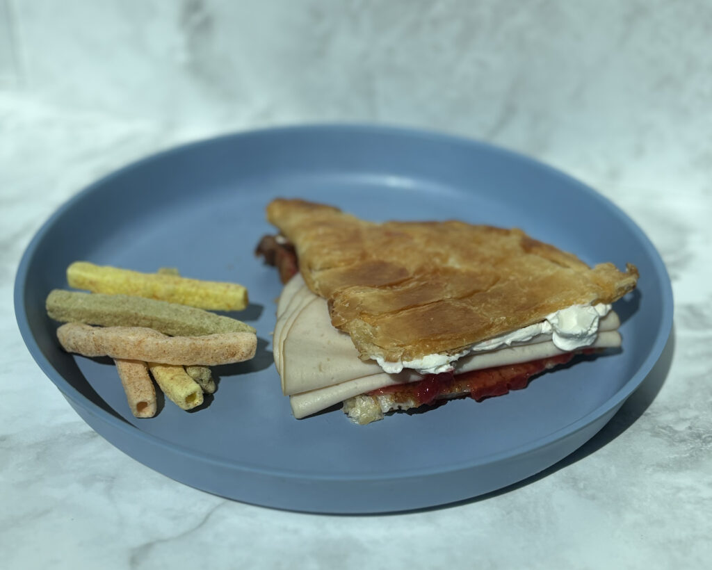 A picture of a turkey, cream cheese, and jelly sandwich on croissant, with veggie sticks on the side.