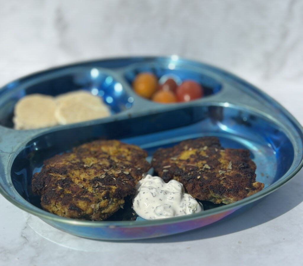 Two meat and potato patties, known as kotlet, appear on a plate with cucumber yogurt; pita and tomato are plated in the background.