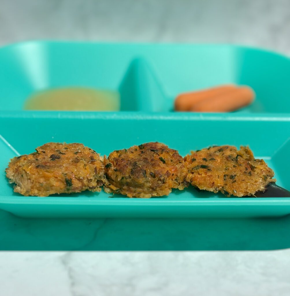 An infant feeding plate foregrounded with three salmon and sweet potato cakes; applesauce and carrots are plated in the background.