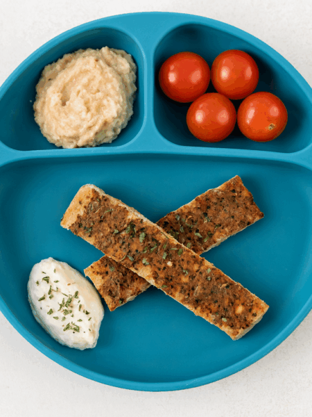 Two sticks of the meat and veggie flatbread known as lahmacun are shown on a plate, sided by herbed yogurt, tomatoes, and mashed potatoes.