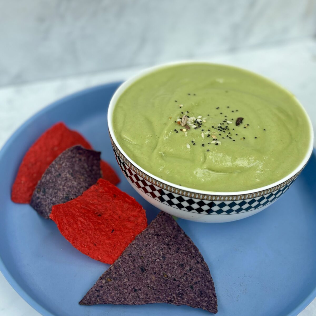 A bowl of pea, avocado, cucumber, and ricotta soup is atop a blue plate; the plate contains tortilla chips of various colors.