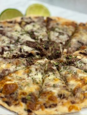 Close-up of a small pizza with black beans and zucchini as the sauce; browned cheese covers it. Limes are provided on the side.