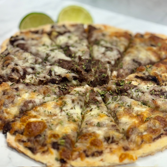 Close-up of a small pizza with black beans and zucchini as the sauce; browned cheese covers it. Limes are provided on the side.