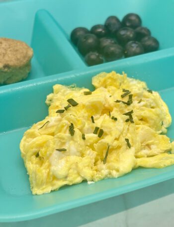 Scrambled eggs foreground a breakfast plate that also contains a muffin and blueberris.