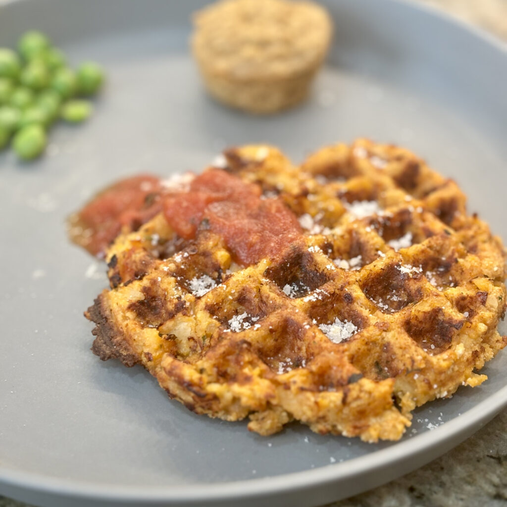 A closeup of a waffle made of chicken, topped with tomato sauce and parmesan cheese.