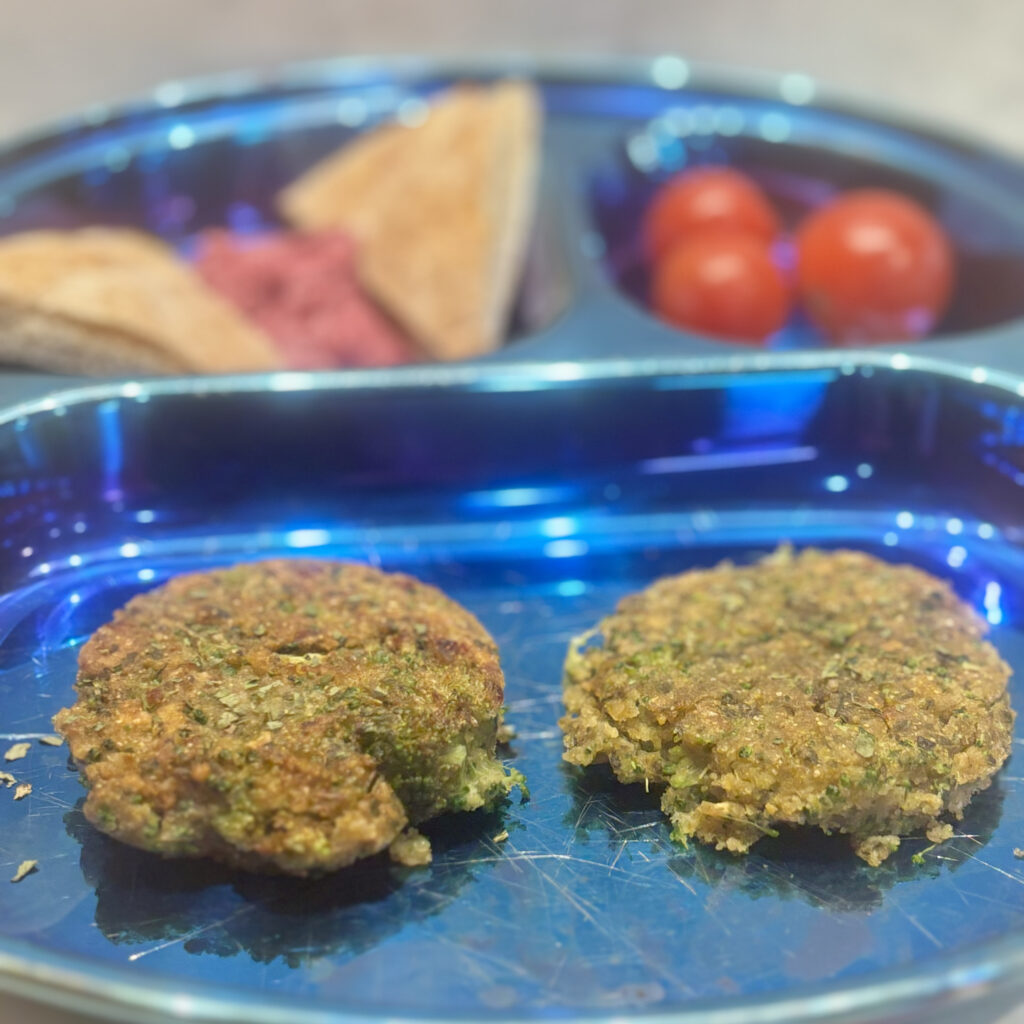 Two falafel patties stuffed with broccoli front a plate that also includes beet hummus and tomatoes.