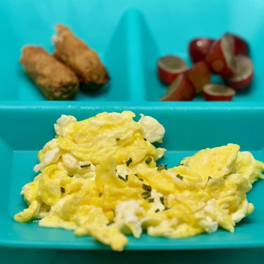 A tussle of scrambled eggs is plated with turkey sausage links and cut grapes.