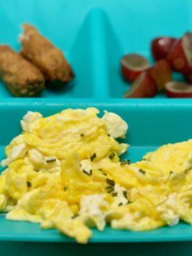 A tussle of scrambled eggs is plated with turkey sausage links and cut grapes.