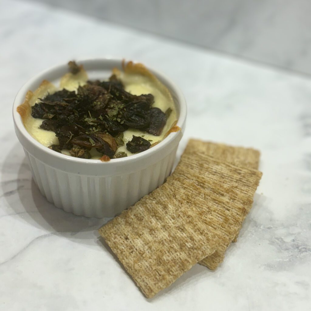 A ramekin is filled with melted cheese and topped with crispy mushrooms; wheat crackers rim the bowl.