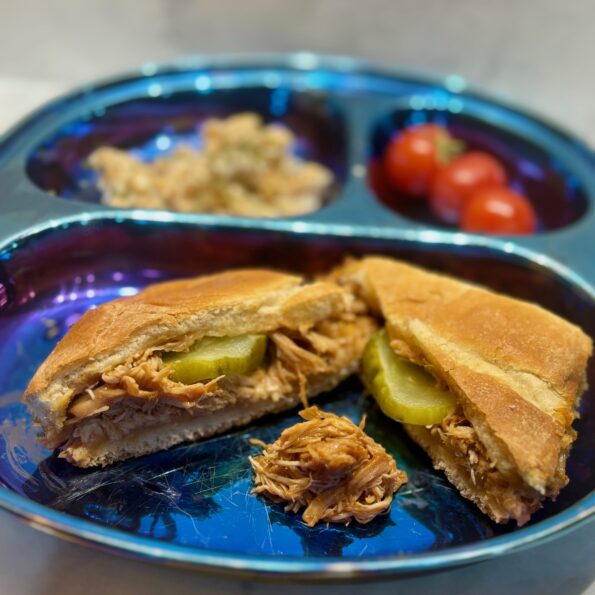 Two halves of a barbecue pulled chicken sandwich, each with a pickle slice inside, are displayed on a plate; a small mound of pulled chicken sits between them.