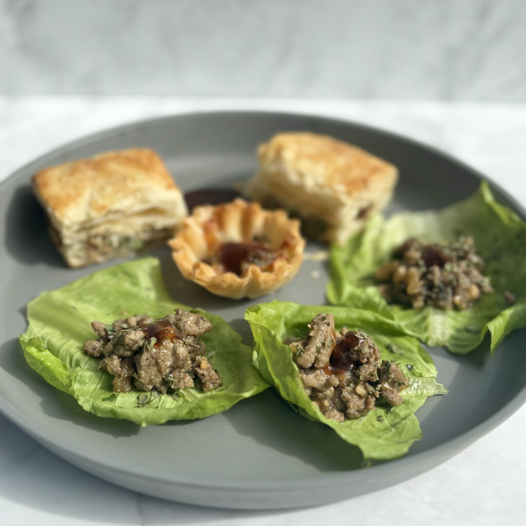 A plate contains three lettuce cups filled with turkey, cashew, and mushrooms; in the background, the same filling is presented in puff pastry and pastry shells