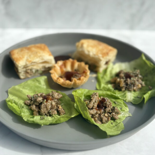 A plate contains three lettuce cups filled with turkey, cashew, and mushrooms; in the background, the same filling is presented in puff pastry and pastry shells