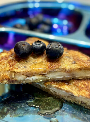 Two halves of a peanut butter and banana-stuffed french toast sandwich are stacked on top of one another, with blueberries placed all over.