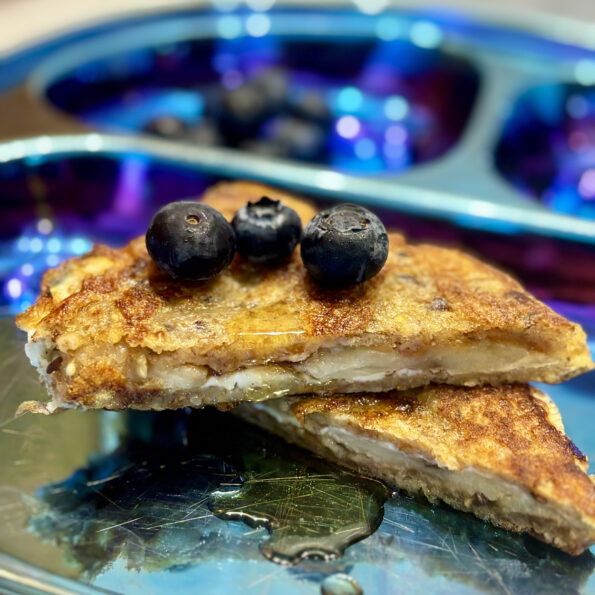 Two halves of a peanut butter and banana-stuffed french toast sandwich are stacked on top of one another, with blueberries placed all over.