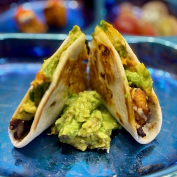 A griddled tortilla filled with beans, avocado, plantains, and cheese is split in half and plated standing up, a pile of guacamole between each half.