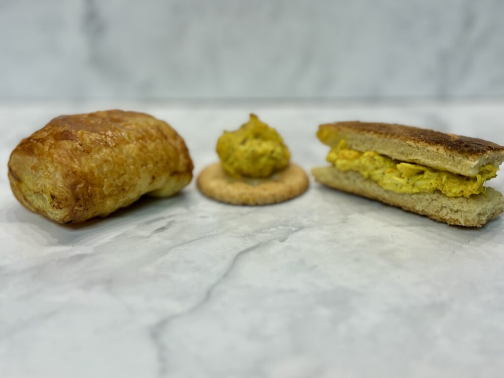 Three forms of chicken salad are presented on a counter: at left, a puff pastry encases the salad; at center, the chicken salad is presented on top of a cracker; at right, it is sandwiched between two slices of toast.