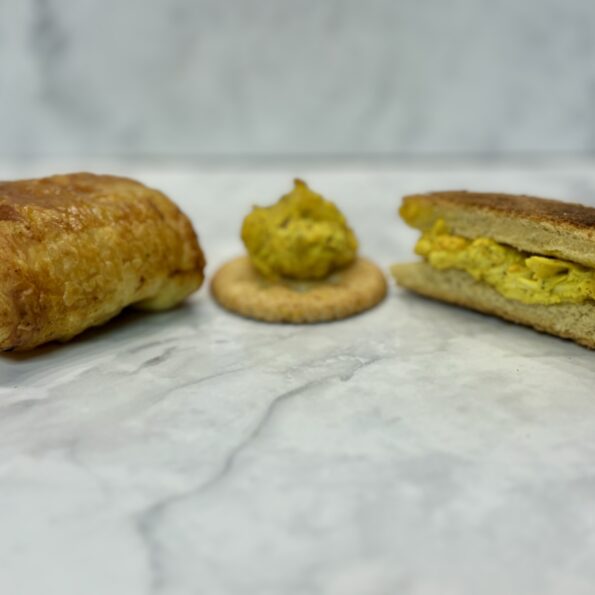 Three forms of chicken salad are presented on a counter: at left, a puff pastry encases the salad; at center, the chicken salad is presented on top of a cracker; at right, it is sandwiched between two slices of toast.