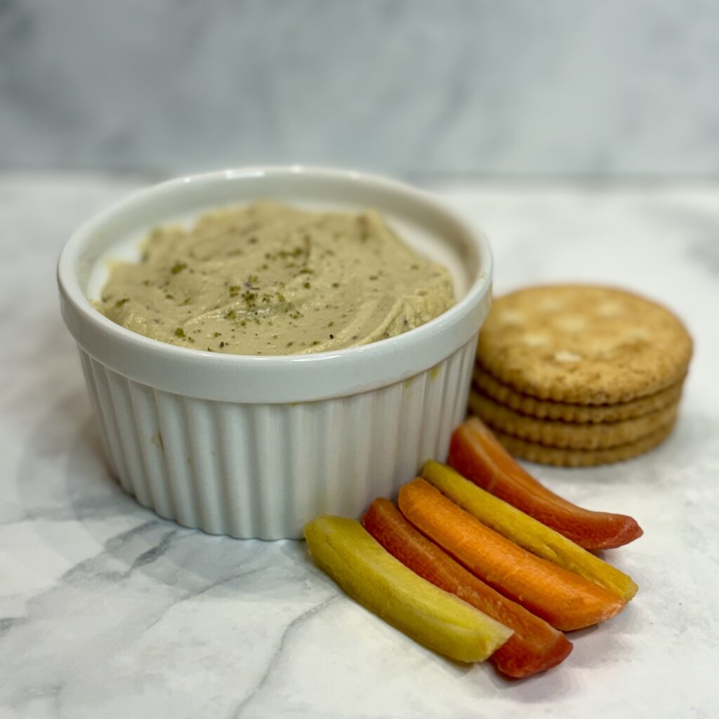 A small bowl of lentil and cauliflower hummus is sided with rainbow carrots and a small stack of crackers.