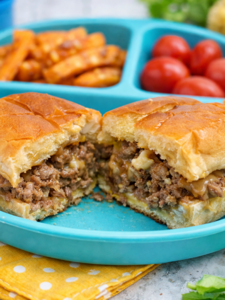 A chopped cheese sandwich, somewhat like a toddler cheeseburger, is shown split in half and served on a child's plate, with fries and tomatoes on the side.