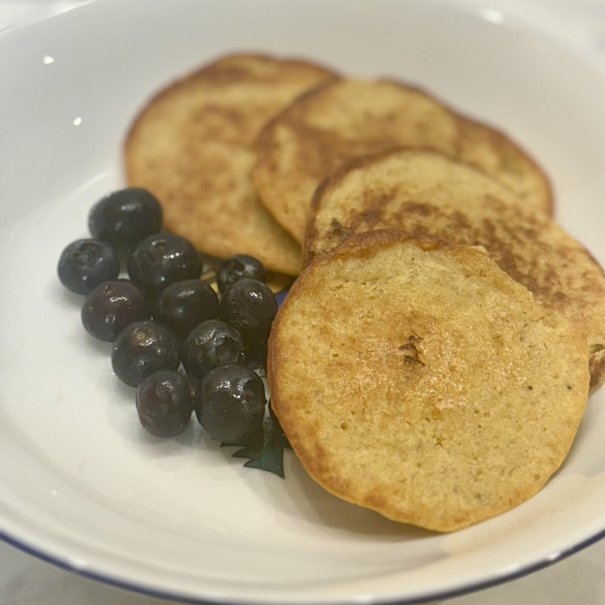 A pile of mini banana-yogurt pancakes is strewn about a plate, sided by blueberries.