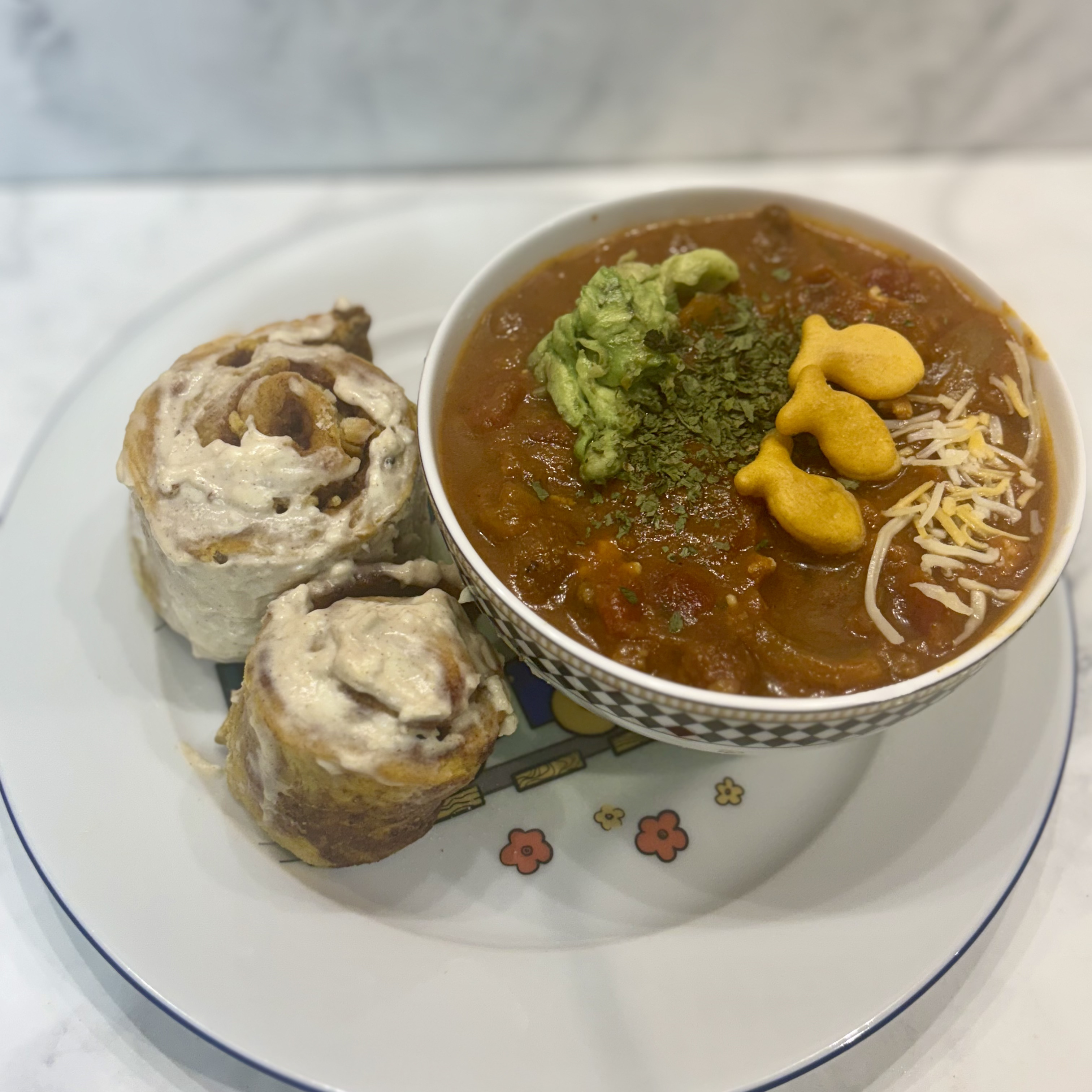 A bowl of toddler chili is topped with avocado, cilantro, cheese, and crackers; on its side are two small pumpkin cinnamon rolls.