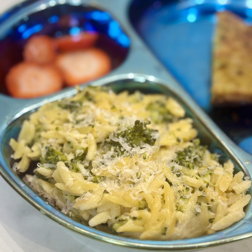 A pile of cheesy orzo with broccoli lines the side of a toddler plate.