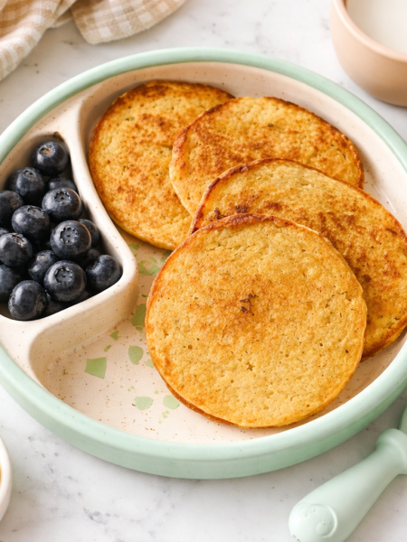 A stack of banana pancakes rest on a plate, alongside blueberries.