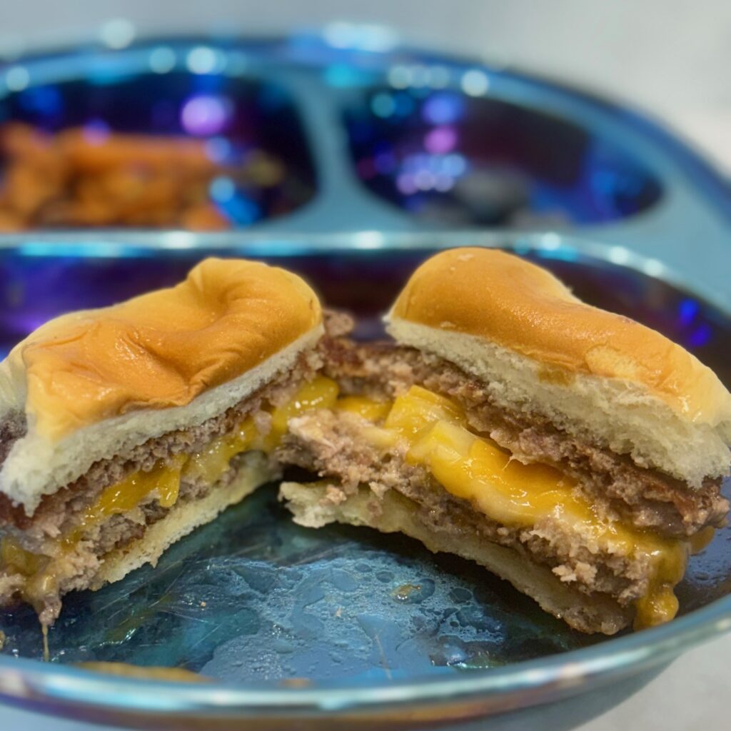 A toddler cheeseburger in the style of a juicy lucy is shown cut in half, with cheese oozing out of the center of the patty.