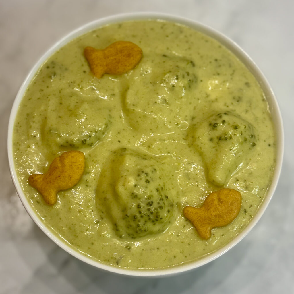 Overhead picture of a bowl of broccoli cheese soup made for kids; broccoli florets and goldfish dot the top.