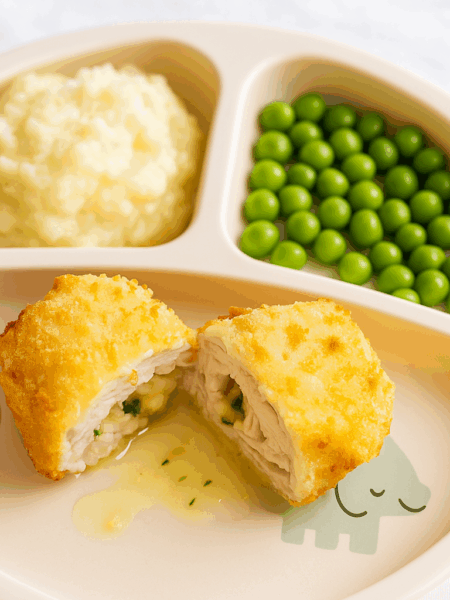 Chicken kiev for kids is shown split, with its butter sauce and herb filling at center; peas and mashed potatoes are included on the plate.