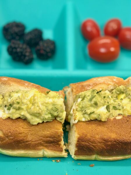 A toddler egg salad sandwich recipe, featuring avocado and tahini, is shown stuffed into a split-top bun.