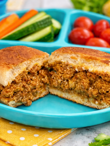 A sloppy joes for toddlers sandwich is shown split open to reveal the meaty insides; the plate also contains sides of crunchy raw vegetables.