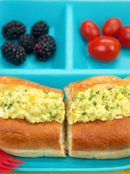 A split-top bun filled with an egg salad sandwich for toddlers is shown on a plate that also includes berries and tomatoes.