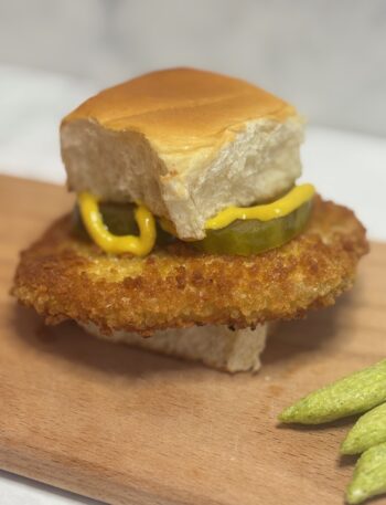 A breaded pork tenderloin fit for a toddler is presented on a hawaiian roll and dressed with mustard and pickles