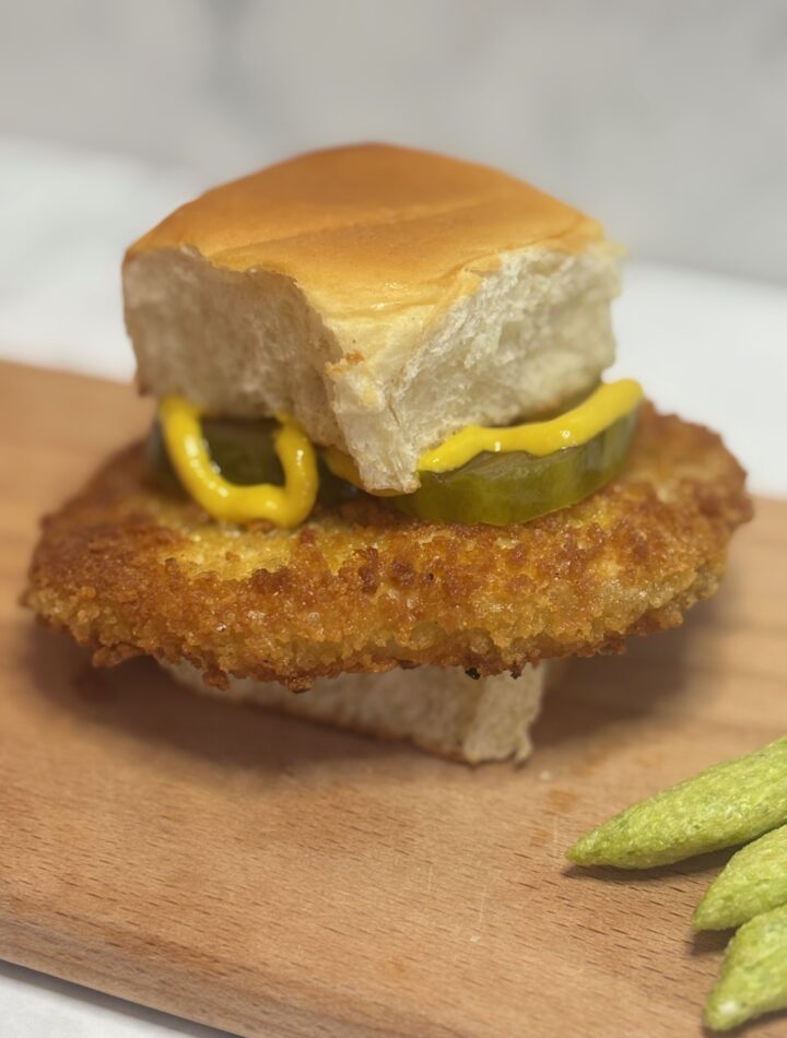 A breaded pork tenderloin fit for a toddler is presented on a hawaiian roll and dressed with mustard and pickles