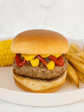 A toddler burger, featuring a toasted mayo bun, smashed patty, and all the condiments; plate also includes corn and fries.
