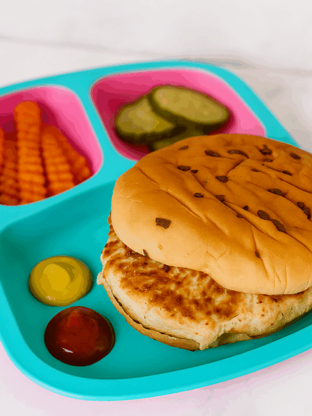 A toddler turkey burger is shown here on a bun, flanked by condiments and sided with sweet potato fries and pickles.