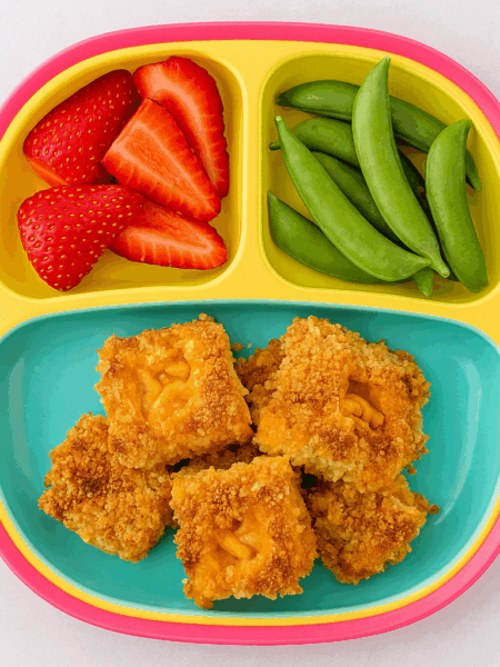air fried mac and cheese bites are stacked on a plate alongside fruit and veggies.