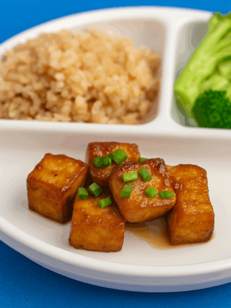 Cubes of tofu, cooked and sauced like orange chicken, are displayed on a plate that also includes brown rice and broccoli.