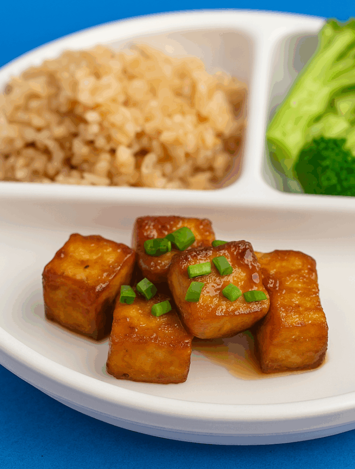 Cubes of tofu, cooked and sauced like orange chicken, are displayed on a plate that also includes brown rice and broccoli.