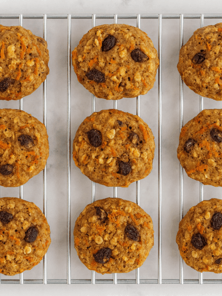 Rows of carrot oatmeal cookies for toddlers are shown resting on a cooling rack, with raisins, carrot strands, and nuts visible.