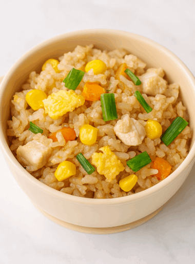 A small bowl is filled with vegetable fried rice for toddlers, with bits of egg, corn, carrot, and green onion visible at top.