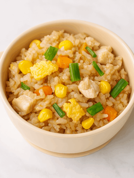 A small bowl is filled with vegetable fried rice for toddlers, with bits of egg, corn, carrot, and green onion visible at top.