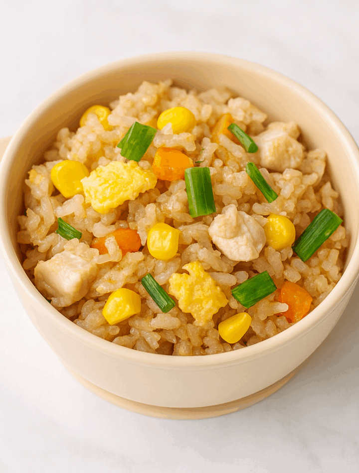 A small bowl is filled with vegetable fried rice for toddlers, with bits of egg, corn, carrot, and green onion visible at top.