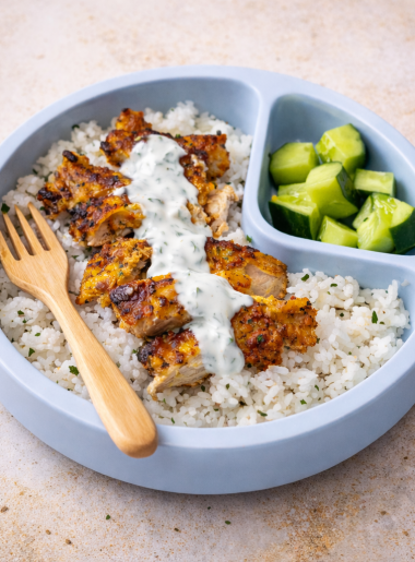 A child's plate features slices of chicken tikka for kids that are covered in a ranch yogurt sauce; the chicken sits atop rice and is sided by cut cucumber.