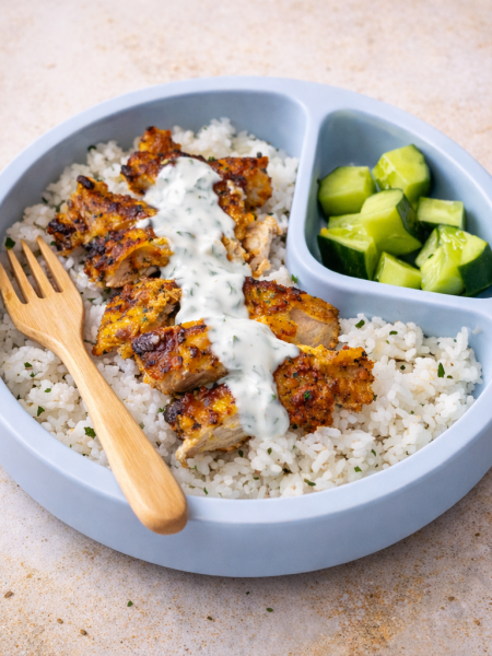A child's plate features slices of chicken tikka for kids that are covered in a ranch yogurt sauce; the chicken sits atop rice and is sided by cut cucumber.