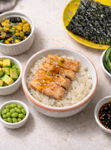 A salmon and rice bowl for toddlers is shown surrounded by various toppings: broccoli and corn salad, seaweed snacks, pickles, soy dressing, edamame beans, and avocado cubes.